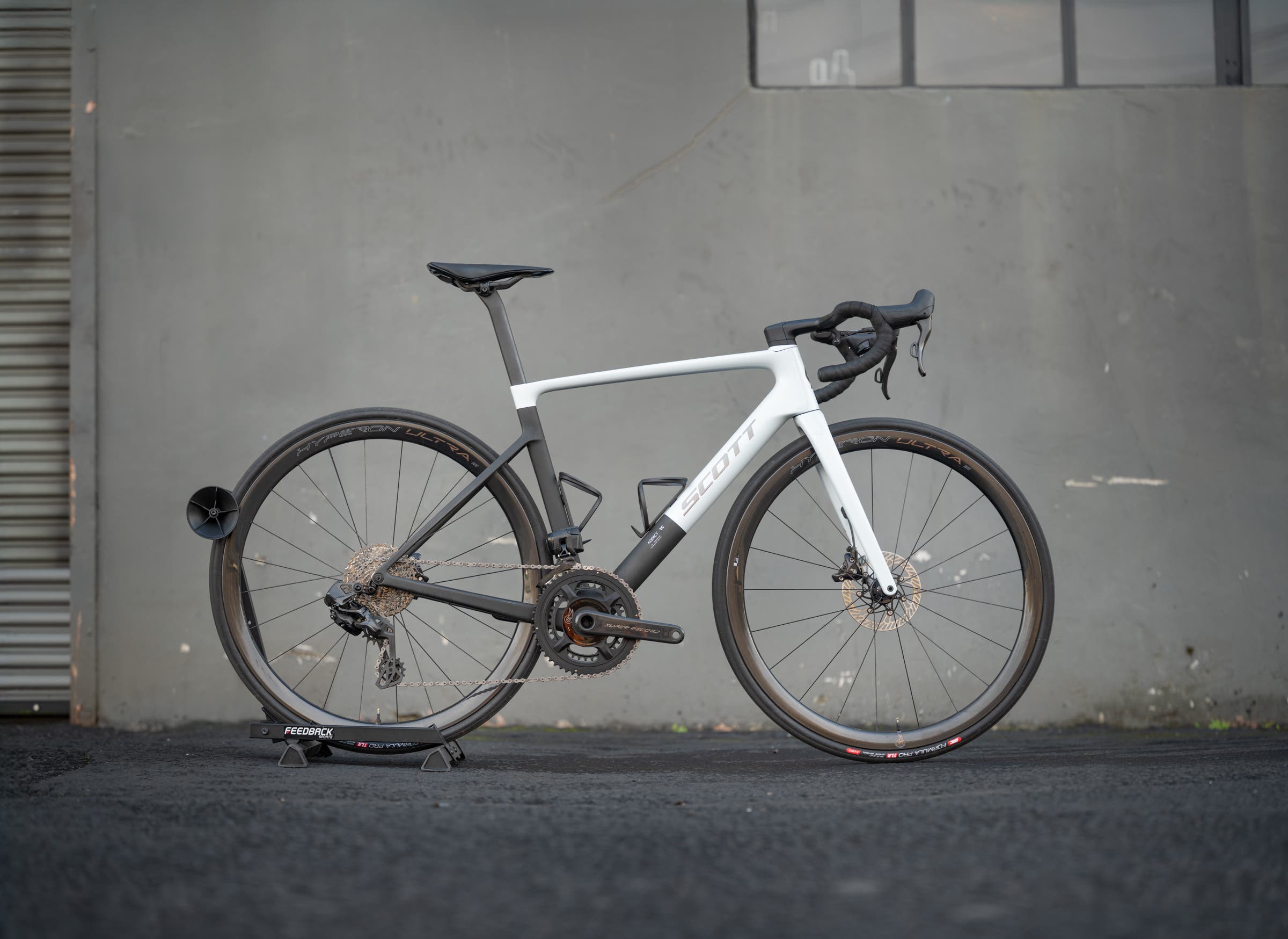 Profile view of a Scott Addict RC road bike equipped with Super Record 13 groupset and Campagnolo Hyperon Ultra wheels against a dark grey industrial wall